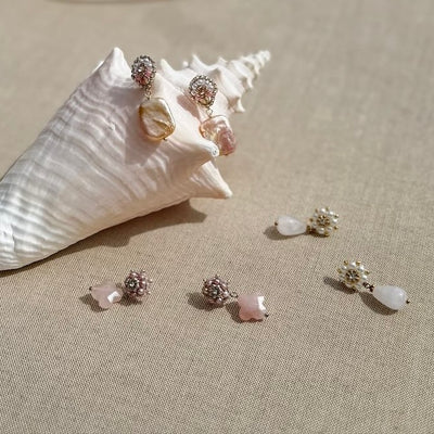 Maschalinas Natural Quad Pearl Ohrringe mit verschiedenen Perlen und Edelsteinen werden auf einer strukturierten Oberfläche mit Muschelschalenhintergrund präsentiert. Diese Designer-Ohrringe strahlen mit ihren glänzenden Perlen in zarten Rosa- und Weißtönen Eleganz und Schlichtheit aus.